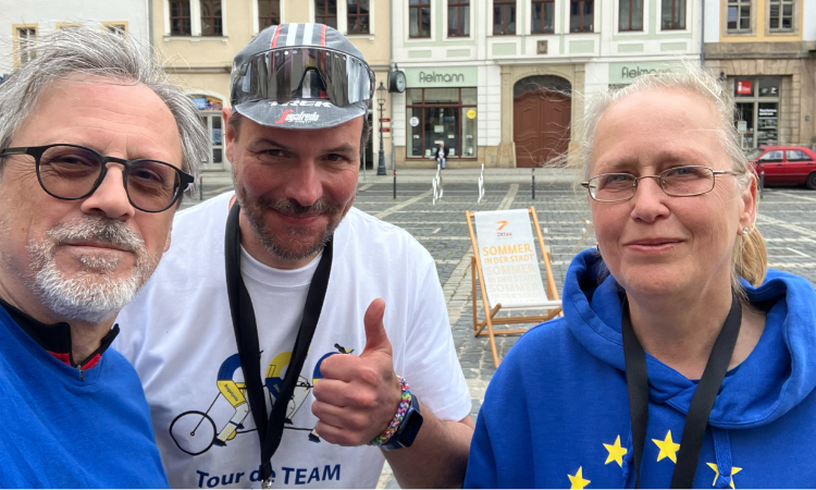 Drei EP-Mitarbeitende, zwei in Europa-Farben und einer mit Radfahr-Kappe und -Brille, lächeln in Zittau in die Kamera. 