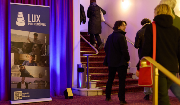 Publikum geht die Treppen zum Filmsaal hinauf. Neben der Treppe steht ein Roll up, das die nominierten LUX-Filme zeigt.